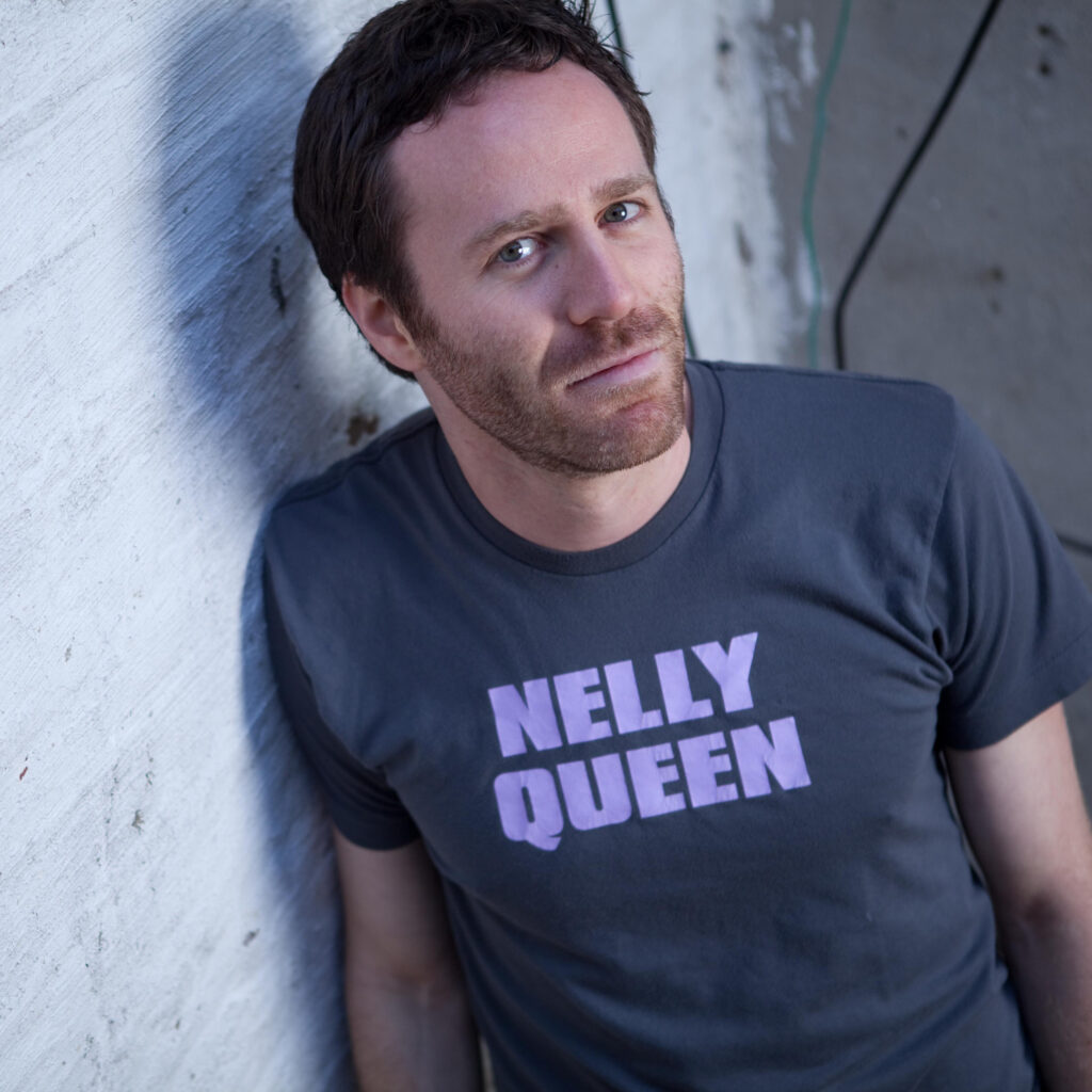 Headshot of Jake Goodman looking at the camera and wearing a t-shirt that says "Nelly Queen"