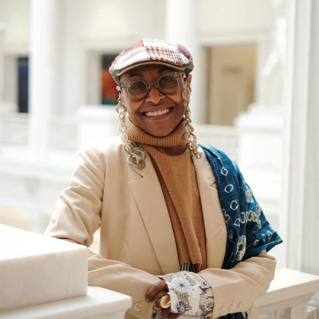 yvette shipman headshot - yvette is smiling toward the camera, and wearing a flat hat, earth toned shirt and jacket, an indigo scarf over one shoulder, and glasses. She looks SHARP!